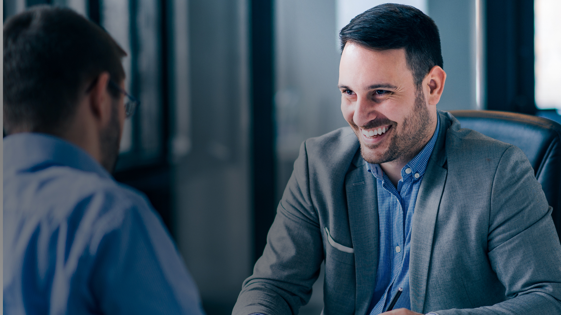 A smiling man in a suit sits across a table from another person in an office setting during a meeting.